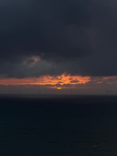 The image captures a dramatic sunrise or sunset over the ocean, showcasing an offshore wind farm. The sky is dominated by a sweeping band of vibrant orange and yellow clouds, horizontally bisecting the frame. This luminous strip, suggesting the sun's low position, is bordered by heavy, dark grey and deep blue clouds above, creating a stark contrast. Below the fiery horizon, the dark, calm surface of the ocean extends into the foreground.
Silhouetted against the brilliant light and stretching across the water are numerous wind turbines. Their distinct shapes, including the central tower and the blades, are clearly visible as dark forms against the glowing sky. The turbines appear to be arranged in a line, emphasizing the scale of the offshore wind farm.
The time of day is clearly either sunrise or sunset, indicated by the intense colors of the sky and the low angle of the light. The weather appears partly cloudy, with a clear horizon allowing the sun's rays to illuminate the cloud band. The scene conveys a sense of serene power, blending natural beauty with human engineering for renewable energy. The location is an offshore wind farm near Fécamp, France. No visible text is present in the image.
