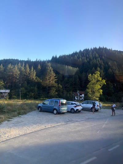 This media file shows a sunny day scene in Eleshnitsa, Bulgaria, likely during the late afternoon given the long shadows. The image is oriented vertically.
The main subjects are several parked cars along a paved road. These include a blue minivan, a white hatchback, and another white car. Two people are visible near the cars; one person is standing by the road, and another appears to be near one of the white cars. Behind the road, a dense forest of evergreen trees covers a hillside under a clear blue sky. A small building with a reddish roof is nestled amongst the trees in the background. The road itself appears to have a gravel shoulder on one side, transitioning to pavement.
The scene suggests a pause or stop during travel, with people interacting with their vehicles in a natural, forested environment. The lighting indicates a bright, clear day with warm sunlight. No visible text is discernable in the image.