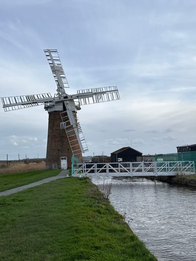 A landscape image taken in the North Norfolk region of the United Kingdom displays a large brick tower windmill as its central subject. The windmill features a white cap and four white sails, each equipped with numerous wooden slats, some of which appear damaged or absent. A white door is visible at the base of the mill's brick tower, with a single rectangular window positioned above it. To the right of the windmill, a narrow waterway with a rippled, dark grey surface flows horizontally across the frame. A white metal pedestrian bridge, characterized by a truss design, spans this waterway. On the far bank, beyond the bridge, a small, dark-colored wooden building, possibly a shed or boathouse, is visible, partially enclosed by green construction fencing. Another partially visible structure is situated further to the right. The foreground consists of green grass, with a narrow, light-colored path running alongside the waterway towards the windmill on the left bank. Tall, dry reeds or grasses extend into the distant background to the left of the windmill, where power lines and poles are also discernible. The sky is overcast, presenting a light grey tone with visible cloud formations. Two small green cone-shaped markers are placed near the entrance of the bridge on the near bank. No individuals are present in the scene. The overall environment is flat and open, indicative of fenland terrain, under overcast weather conditions.