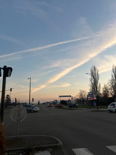 The image depicts an urban road intersection in Vendenheim, France, under a sky exhibiting a light blue to orange-pink gradient, indicative of dusk or dawn. Multiple white contrails traverse the sky. A prominent Castorama retail sign is visible in the mid-distance.

In the immediate foreground, to the left, a dark pedestrian traffic light is mounted on a pole. A blank circular road sign, oriented away from the viewer, stands on a median strip where minimal vegetation is visible alongside patches of what appears to be residual snow or frost.

Vehicular traffic is present. A dark grey sedan is captured in the midground, executing a left turn at the intersection. Further down the primary road, an orange hatchback proceeds away from the viewer. On the right side of the frame, near a multi-directional street sign displaying "LAMPERTHEIM" and a large, dark electronic display board, several vehicles are stationary or parked, including a white hatchback. Bare deciduous trees line the roadsides and the distant horizon. Streetlights, some equipped with security cameras, are spaced along the road. The road surfaces are asphalt, marked with lane delineations. No individuals are visible.