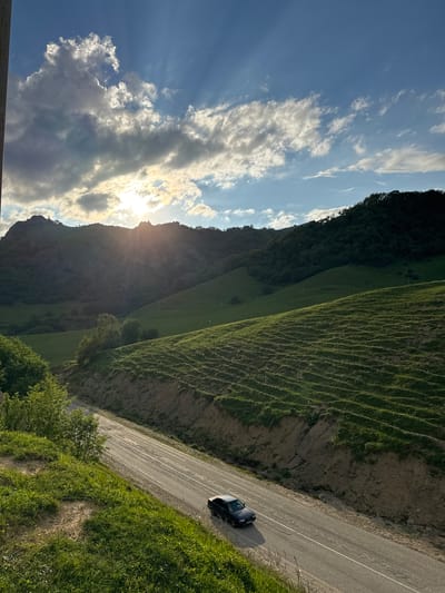 The image depicts a scenic view of rolling green hills and mountains under a bright, partly cloudy sky with the sun setting. A paved road runs diagonally across the foreground, with a dark-colored car driving on it. The hills on either side of the road are covered in lush green grass, with some areas showing terraced patterns. In the background, a large mountain range is visible, with dense green trees covering its slopes. The sun is low on the horizon, casting long shadows and creating a dramatic effect with sunbeams breaking through the clouds. The overall impression is one of natural beauty and tranquility. The location is identified as Nizhnyaya Mara, Russia.