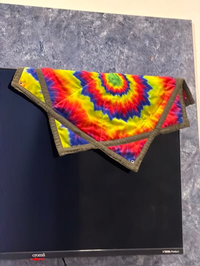 A tie-dye blanket with red, yellow, blue, and green colors is draped over the top of a black television screen. The blanket has a star-like pattern radiating from the center and is bordered with a darker fabric. The television is mounted on a wall with a textured gray finish. The bottom edge of the television displays a silver bar with the brand name "Croma" in red text on the left and "A TATA Product" in white text on the right. The scene appears to be indoors, likely in a residential or commercial setting.