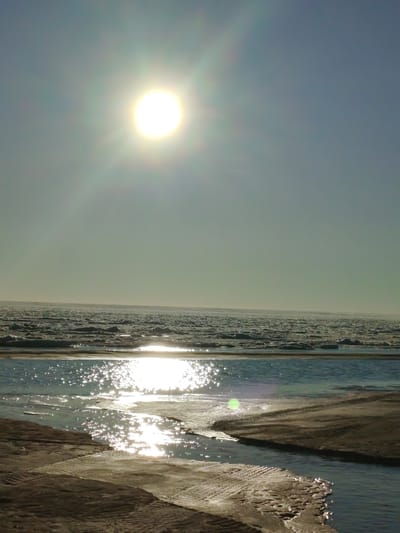 A bright sun shines in the upper left corner of the image, its rays creating a lens flare that extends across the sky. Below the sun, the sky is a pale blue that transitions to a light gray near the horizon. The horizon is marked by a vast expanse of ice floes, broken and uneven, stretching as far as the eye can see. The sun's reflection glints off the water in the foreground, where a narrow stream of water flows between sandy banks. The banks appear to be a mix of sand and ice, with tire tracks visible in the sand. The water is dark blue and reflects the bright sunlight in shimmering patches. The overall impression is of a cold, desolate landscape under a bright, but not necessarily warm, sun.