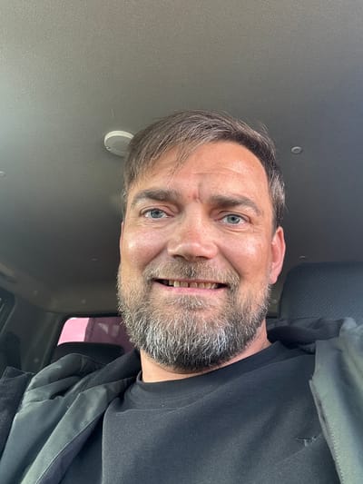 A medium-shot selfie is taken from inside a vehicle. The subject, a middle-aged Caucasian male with brown hair and a full grey beard, is looking directly at the camera. He is wearing a black t-shirt and a black jacket. The interior of the vehicle, including the ceiling and a car seat, is visible in the background. The scene appears to be located in Parma, Russia.