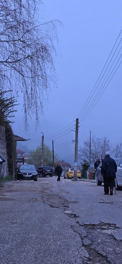 The image depicts a street scene in Breze, Bulgaria. Two people are walking on a cracked asphalt road. A parked silver car and a yellow car, as well as other vehicles, are visible. Overhead, a telephone pole with five wires stretches across the sky. A bare tree with thin branches is in the foreground. The sky is overcast.