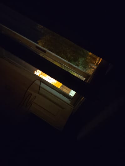 An outdoor night view through a window frame shows illuminated building facades and trees. The left window pane reveals a portion of a light-colored door with dark line drawings on its surface, possibly graffiti or a natural feature like condensation. To the right, another window pane shows a darker scene with blurred yellow lights suggesting streetlights or interior illumination of distant buildings, and dark green foliage from trees or bushes. The scene appears to be from an urban setting, potentially in Arad, Romania, given the context provided. The image is dark, suggesting it was taken at night.