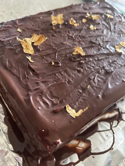 A close-up, overhead shot shows a rectangular chocolate cake with a glossy dark chocolate frosting. The frosting is unevenly applied, with visible ridges and swirls, suggesting it was recently poured or spread. Scattered across the top of the cake are irregular flakes of gold leaf, reflecting the light. The cake is placed on crumpled aluminum foil, with drips of chocolate extending over the sides and onto the foil. The background is a blurred, light-colored surface, possibly a countertop. No people are visible in the image. The context of Richmond Hill, Canada, is not depicted in the visual elements of the photograph.