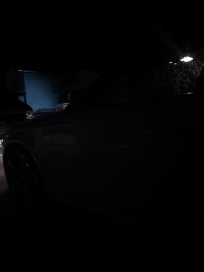 An image taken at night shows a dark, partially illuminated scene in Dolni Tsibar, Bulgaria. A silver sedan is parked in what appears to be a residential area or driveway. The car's side and wheel are visible on the left, with the majority of the vehicle obscured by darkness. In the background, a building is dimly lit with a blueish light, and there are glimpses of what might be other vehicles or structures. The upper right portion of the image shows a window with rain streaks and a bright overhead light. The overall atmosphere is dark and moody, with limited visibility.