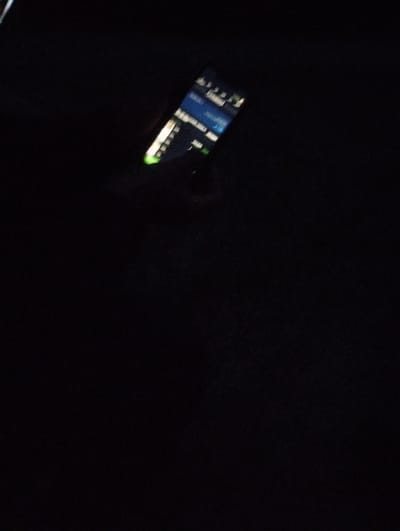 The media file shows a close-up, low-light shot of a smartphone screen. The phone is angled, and the screen displays a user interface with text and graphics, suggesting an application or operating system is active. The surrounding environment is very dark, indicating that the image was likely taken at night or in a dimly lit interior. The phone screen is the primary light source in the image. Some text is partially visible on the screen, including what appears to be list items with accompanying icons or statuses, and some text that might be headings or labels, but is too blurry to decipher clearly. There are also some green elements visible on the right side of the screen, possibly indicating status indicators or interactive elements. No people or clear setting details beyond the darkness are discernible. The context provided suggests the location is Brashlen, Bulgaria.