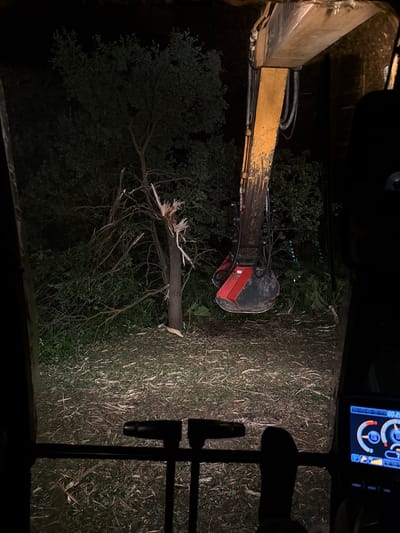 An outdoor night scene shows the view from inside a construction vehicle, likely an excavator, looking towards a damaged tree. The excavator's yellow arm and red cutting attachment are prominent in the foreground, extending into the dark, wooded area. The tree in the mid-ground is broken and splintered near its top, with branches scattered around its base and on the ground. The ground is covered in debris, including wood chips and broken branches. The background consists of dense foliage and dark trees. A digital display is visible in the lower right corner of the frame, showing graphical readouts. The image appears to be taken in Kutepovo, Russia, a location that suggests a rural or natural environment.