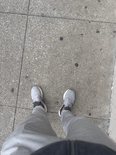 A person's legs and feet are visible in the image, standing on a concrete sidewalk. The person is wearing grey sweatpants and grey athletic shoes with white soles and black accents. The sidewalk is made of concrete slabs with visible cracks and dark spots, possibly from dirt or stains. The lighting suggests it is daytime. The scene appears to be an outdoor urban environment, consistent with a sidewalk in San Francisco, United States of America.