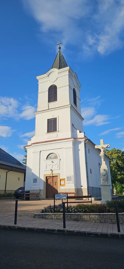 The image shows the exterior of a white church with a dark, peaked steeple topped with a cross. The church has arched windows with louvers and a date, "1833," inscribed above the entrance. A large wooden door with a cross design is central to the facade. To the right of the church entrance is a statue of Jesus on the cross with another smaller figure at its base, dated "1898." There are two wooden benches outside the church and a sign that reads "KOSSUTH LAJOS UTCA." A parked car is visible to the left of the church, alongside a building with solar panels. The scene is set against a bright blue sky with scattered clouds. The location is identified as Balatongyörök, Hungary.