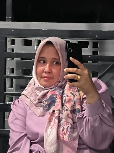 A woman wearing a light purple hijab and a matching purple dress is taking a selfie with a black smartphone. She is looking to her right, away from the camera. The background appears to be an outdoor setting, possibly a patio or balcony, with a dark, textured wall with geometric cutouts. The lighting suggests it is evening or nighttime. The woman is wearing a gold ring on her right hand.