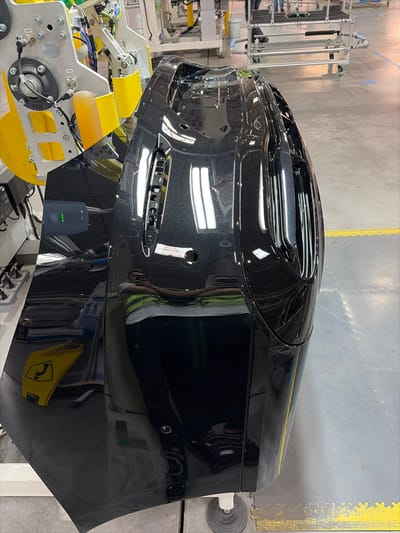An industrial scene is depicted within a manufacturing facility located in Bad Rappenau, Germany. The primary subject is a highly reflective, glossy black automotive body panel, possibly a bumper or fender, featuring integrated vents and grilles. This panel is held by a silver-gray robotic arm equipped with yellow components, visible tubing, and wiring, indicating a stage in automated production or inspection. The immediate environment consists of a light gray industrial floor marked with yellow tactile safety strips. Overhead fluorescent lighting fixtures are prominently reflected on the polished surface of the automotive panel. Reflected on the lower portion of the panel is an adult male, seen holding a smartphone as if taking the photograph. He wears a white short-sleeved shirt, a high-visibility yellow-green safety vest, dark pants, and dark athletic shoes. In the background, other industrial equipment, including a metal trolley with horizontal bars, is faintly visible.