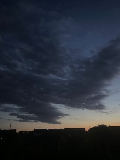 A low-angle, full-frame, vertical shot captures a dusky sky filled with dark, ominous clouds. The sky transitions from a deep navy blue at the top to a soft orange glow near the horizon. Silhouetted against this sky are the rooftops of low-rise buildings, suggesting a residential area. Some windows in the buildings are illuminated, indicating indoor activity. The foreground is entirely dark, obscuring any details of the ground or lower structures. The scene is devoid of any visible people or specific landmarks, making the location in Slobozia, Moldova, an assumption based on prior context rather than explicit visual evidence.