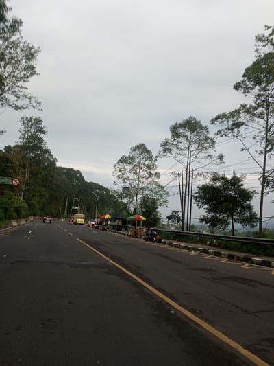 The image shows a roadway on the outskirts of Taunggyi, Myanmar. The road is black asphalt with a single yellow line separating traffic lanes. A guardrail runs along the right side of the road, with trees and foliage visible beyond. Several vehicles, including a red car and a yellow truck, are on the road. There are also several stalls or stands with colorful umbrellas set up on the side of the road. The sky is overcast. A sign reading "JL Raya Puncak" is visible.