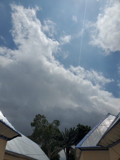 The image depicts a partially cloudy sky over a residential area in Kason, Nigeria. Two corrugated metal roofs of houses are visible in the foreground, one on the left and one on the right, with blue trim. Tall trees, including what appears to be a palm tree, are situated behind the roofs. The sky features a mix of bright white clouds and darker, more ominous storm clouds, suggesting a transition in weather. A distinct sunbeam or lens flare cuts diagonally across the upper right portion of the sky. There are no people or animals visible in the scene.