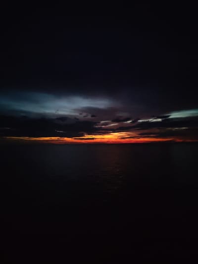 The image shows a dark sky over the sea at dusk. A thin band of orange and red is visible on the horizon where the sun has set. Dark clouds are scattered across the sky, with some lighter gray and blue hues present. The sea is dark and calm, with a slight shimmer on the surface reflecting the last light from the sky. The scene is very dark overall, with the exception of the vibrant sunset colors. There are no people or man-made objects visible in the image.
