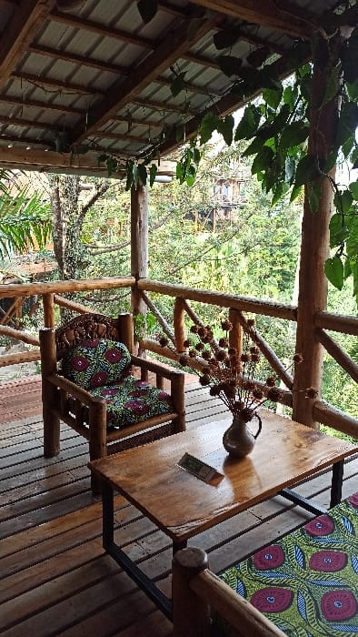A wooden patio or deck with a table, chairs, and greenery overhead. The table is set with a vase of dried flowers and a menu. The chairs are decorated with colorful patterned cushions. In the background, trees and another wooden structure are visible through the open framework of the patio. The scene gives a sense of nature and relaxation.