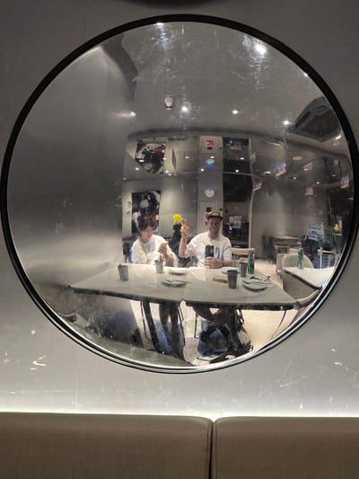 A circular mirror reflects a scene in what appears to be a cafe or restaurant. Two individuals are seated at a table. The person on the left, wearing a white t-shirt, is looking at their phone and holding a yellow object. The person on the right, also in a white t-shirt and wearing a baseball cap, is holding a phone and making a peace sign with their left hand. The table is set with plates, cups, and napkins. In the background of the reflection, other tables and chairs are visible, along with ceiling lights and decorations on the walls. The mirror is mounted on a grey, textured wall, and a brown upholstered bench is visible in the foreground of the main image. The mirror shows signs of being smudged and scratched. The location is Fuyang, China.
