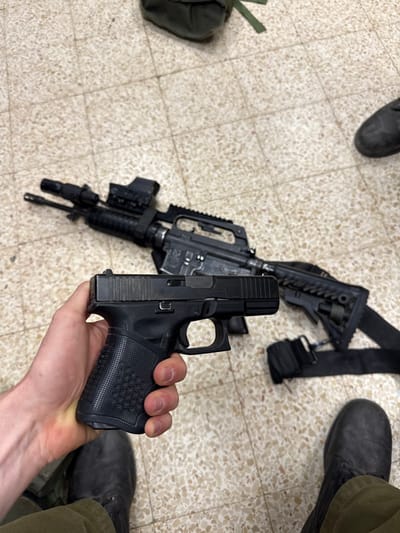 A person is holding a black Glock 19 pistol. The pistol has a black polymer frame and a black metal slide with a textured grip. The person is wearing black tactical boots and olive green pants. A black rifle with a black strap lies on the beige tiled floor. There is a green military backpack on the floor in the upper left corner of the frame. The location is a room in Mazkeret Batya, Israel, based on the coordinates 31.833, 34.82.