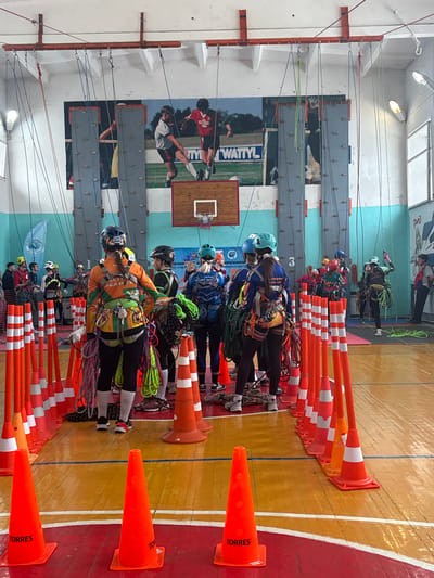 An indoor gymnasium in Tayshet, Russia, is depicted during a rope skills or climbing event. The floor is polished wood with red and yellow sections marked by white and blue lines, and lined extensively by orange plastic traffic cones, many bearing the "TORRES" label. The back wall is painted light blue and white, featuring a large mural of soccer players in action, one in a white jersey and another in a red jersey, with the word "WATTYL" visible. Below this mural, several grey climbing wall panels are mounted. A basketball hoop with a white net is affixed to the back wall. Overhead, an orange metal beam structure is installed across the ceiling, from which numerous colored ropes (blue, green, red, purple) hang downwards.
Approximately 15-20 individuals, primarily young adults, are present in the scene. Most participants are equipped with helmets (various colors including blue, yellow, and black), full-body climbing harnesses adorned with multiple carabiners and gear loops, and athletic clothing. Several individuals in the foreground, facing away from the viewer, are preparing for or engaged in an activity, handling coiled ropes attached to their harnesses. One individual in the center foreground wears an orange and green long-sleeved shirt, black pants, and a dark helmet, with a braided ponytail visible. Other groups of individuals are scattered throughout the gymnasium, observing or interacting near the climbing wall elements. The overall scene suggests a structured training session or competition emphasizing rope access and safety techniques.