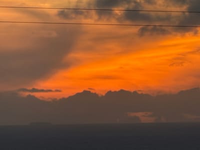 An orange cloud formation dominates the scene, contrasted by darker, gray clouds to the right and above. The orange cloud appears vibrant, illuminated by a source of light. Power lines stretch horizontally across the lower portion, adding a man-made element to the otherwise natural landscape. The sky above the orange cloud transitions into a dark, overcast hue, suggesting a storm or approaching nightfall.