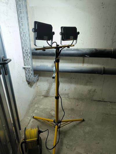 In a room with concrete walls and floor, a yellow tripod work light with two LED lamps is positioned centrally. To the left, a metal cable tray is affixed to the wall, and a silver metal doorframe is visible. In the foreground to the left, a yellow extension cord is coiled on a black reel. Two large gray pipes run horizontally along the wall above the work light. The scene appears to be an unfinished or industrial space.