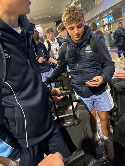 The image depicts a group of individuals, primarily young males, within the interior of O.R. Tambo International Airport (JNB) in Kempton Park, South Africa. The scene appears to be an airport terminal concourse, possibly a check-in or boarding gate area, evidenced by the visible "BRITISH AIRWAYS" and "A59" signage in the background.
In the foreground, a partial view of an individual wearing a dark blue Canterbury-branded half-zip top is visible on the left, with their hand on the handle of a black rolling suitcase. The central and most prominent figure is a young male with light brown, curly hair, dressed in a dark blue hooded sweatshirt beneath a dark blue puffer vest that features a white "POCKLINGTON SCHOOL" logo. He is also wearing light blue shorts with white and orange stripes, white socks, and light-colored sneakers, and carries a dark backpack. This individual is observed holding a small stack of papers or cards in his left hand, looking down at them, while his right hand rests on the extendable handle of a black rolling suitcase.
Behind the central figure, other individuals are visible: a young male on the left, partially obscured, holding a smartphone; a young male in a white polo shirt with a dark emblem and a dark backpack looking towards the right; and a partially visible female further back. In the blurry background, multiple other figures, some with backpacks, are present, including one male with a British Airways-branded bag. The environment features a light-colored tiled floor, bright overhead lighting causing reflections, and airport counters or desks with signs. The group appears to be standing in a queue or waiting area with their luggage.