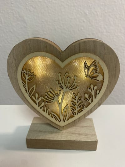 A wooden heart-shaped ornament, illuminated from within by warm LED lights, sits on a white surface. The ornament features a layered design with an outer wooden heart framing an inner heart that is intricately carved with floral motifs and a butterfly. The inner heart's background has a shimmering, textured gold finish. The ornament is supported by a small, rectangular wooden base. The background is a textured white wall. The ornament is located in Sachsenheim, Germany.