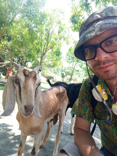 A man wearing a camouflage bucket hat and glasses is taking a selfie with a goat. The man is on the right side of the frame, looking towards the camera. He is wearing a colorful, patterned shirt and a tan hat. A goat with large ears and brown and tan markings is in the foreground, to the left of the man, also looking towards the camera. Behind the goat and to the right of the man, another goat, this one black, is visible. In the background, there are trees, greenery, and two individuals standing. The scene appears to be outdoors, possibly in a rural or park-like setting. The location is identified as Khánh Vĩnh, Vietnam.