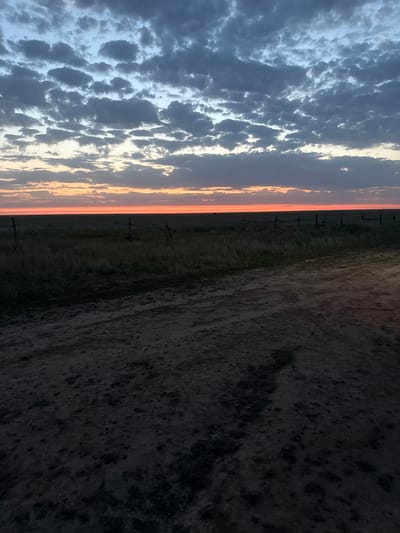 This is a vertical, eye-level photograph of a rural landscape at dusk. The sky is filled with a dramatic display of clouds, transitioning from a pale blue at the top to hues of gray and dark blue lower down. A striking band of bright orange light sits just above the horizon, indicating the last rays of the setting sun.
Below the sky, a flat, open field stretches across the middle ground. This field appears to be covered in dry grass and sparse vegetation. A rustic fence made of wooden posts and wire runs horizontally across the field, partially obscured by the low-lying plants. The ground in the foreground is a rough, dirt road or path, characterized by uneven texture and shadows, suggesting it is unpaved and possibly damp in places. The overall scene conveys a sense of vastness and natural beauty, characteristic of the open steppes of Kazakhstan. The location is Бөрлі ауылдық округі, Kazakhstan.