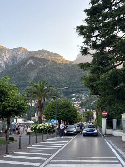 The image shows a street scene in Nobiallo, Italy. In the foreground, a pedestrian crossing spans the asphalt road, with several cars stopped at it. A blue BMW is positioned directly on the crossing, its headlights illuminated. To the left, two pedestrians stand near a parked silver Skoda. Further down the street, other cars and pedestrians are visible. A tall palm tree stands near a lamppost, and to the right, a "no entry" sign is posted next to a fence. In the background, lush green mountains rise, dotted with buildings, and a body of water is visible. The sky is clear and light blue, and the sun casts a warm glow on the mountains.