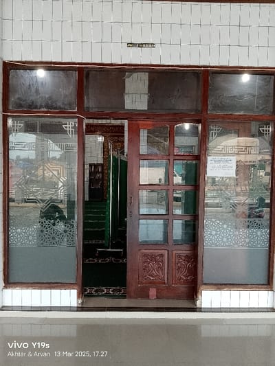 The image shows the entrance to a building at the coordinates -6.44, 108.028 in the city of Kecamatan Gabuswetan, Indonesia. There are three glass doors, two of which are open. One door is closed and appears to be made of wood. A sign above the door reads "ANGGARAN KAS ISLAM". A green rug is visible inside, along with a wooden shelf with books on it. An IV stand is also visible inside the building. The floor outside the building is made of white tiles. The image was taken at 17:27 on March 13, 2023.