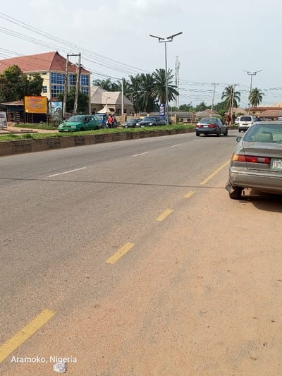 The image shows a street in Ado Ekiti, Nigeria, located at coordinates (latitude: 7.656, longitude: 5.23). There are several vehicles parked and driving along the street. The vehicles include a green car, a silver car, a light gray car, and a dark gray car. There is also a motorcycle with two people on it parked on the side of the road. There are several buildings on the left side of the street, with some trees and power lines in the background. The street is paved with asphalt and has yellow lines painted on it. The sky is clear and there are no clouds. There are palm trees on the right side of the road and there is one streetlight on the left side of the road. The image is taken from a point of view slightly elevated above the street. The image is taken from a point of view slightly elevated above the street. The image is in focus and well-lit. There are no people visible. The image is clear and the resolution is high. The image is of good quality. The image is daytime. There is no sign of rain or precipitation. There is no signage visible. The image is not artistic and does not portray any particular emotion. The image is objective and descriptive. The image does not depict any violence.