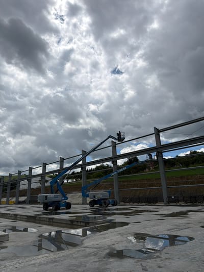 An outdoor construction scene under a cloudy sky is depicted in Skrzydlna, Poland. The foreground shows a wet, concrete surface with puddles reflecting the environment. Two blue boom lifts are positioned on the concrete. One lift, closer to the viewer, has the model "Genie S-85" visible on its base. The second boom lift, further in the background, is extended upwards, with a person wearing an orange vest in the elevated basket. The middle ground features the skeletal structure of a building under construction, composed of large steel beams and vertical supports. Behind the structure, a sloped, grassy hillside is visible, with trees at the top and some buildings in the distance. The sky is dominated by heavy, grey clouds with patches of lighter sky.