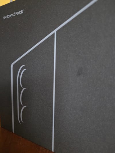 An angled, close-up shot of a dark gray textured box with the white text "Galaxy Z Fold7" in the upper left corner. A thin white border outlines a rectangular section on the left side of the box, which contains three curved indentations that resemble camera lenses. The box rests on a wooden surface. The image is taken from a slightly elevated perspective.