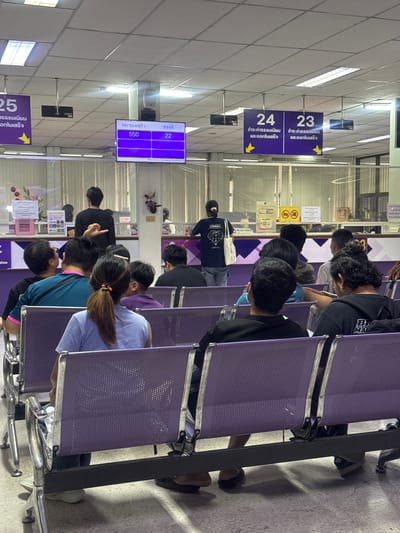 The image depicts the waiting area of the Mae Hia Town Municipality, Thailand. The scene is indoors, under fluorescent lighting, with a tiled floor and a suspended ceiling. In the foreground, a row of purple seats faces a counter area behind a clear partition. Several individuals are seated, waiting, with their backs mostly to the camera. One person in the center is standing and facing away from the camera, gesturing with their left hand.

Above the counter, a digital display board shows queue numbers. The left side of the board reads "หมายเลขคิว" (Queue Number) and the right side reads "ช่องที่" (Channel/Counter). The displayed numbers are 550 (Channel 22), 277 (Channel 23), and 275 (Channel 24). To the left and right of this display are directional signs indicating counter numbers 24 and 23 respectively, with yellow arrows pointing downwards. These signs also have Thai text which appears to relate to fees and completion of services.

The counter area itself has several workstations, separated by clear panels. Behind the counter, some employees are visible, and there are signs and paperwork on display. A few decorative floral arrangements are placed on the counter.

The overall impression is of a government service office or administrative building where citizens are waiting to be served. The presence of the digital queue system and directional signs suggests an organized process for managing public traffic.