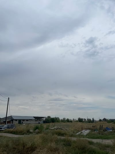 The image depicts an overcast sky over a rural landscape in Kénen Ázіrbayev aýyly, Kazakhstan. In the foreground, dry, tall grass covers the ground, with a dirt road winding through it. A few scattered concrete slabs and debris are visible in the grass. In the midground, a series of low-lying buildings with corrugated metal roofs are present, appearing to be agricultural or storage structures. A dark-colored SUV is parked to the left of the buildings. A wooden utility pole with wires stretches up towards the sky on the far left. The background shows a line of trees and distant structures, including what appear to be power lines and a residential house with a dark roof. The overall impression is one of a quiet, somewhat unkempt rural setting under a heavy, cloudy sky.