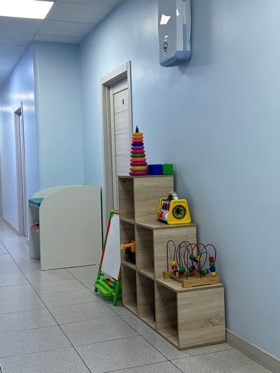 The image depicts a room with light blue walls and a grey tiled floor. On the left side of the image, a white and blue appliance, possibly a humidifier or air purifier, is mounted on the wall. In the center, a wooden shelving unit is filled with various children's toys, including a bead maze, colorful stacking rings, and a yellow toy house. To the right of the shelving unit, a green toy dustpan and broom set is visible. The overall setting suggests a child-friendly environment, perhaps a playroom or a classroom. The lighting indicates it is daytime. No people are visible in the image.