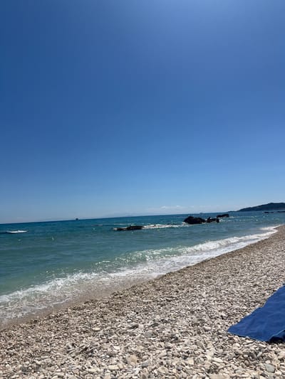 The image shows a pebble beach in Elaties Municipal Unit, Greece. The sea is a vibrant turquoise, with small waves lapping at the shore. Rocks are visible in the water, and a ship can be seen in the distance on the horizon. The sky is clear and blue. In the foreground, a blue towel is laid out on the pebbles. The scene depicts a tranquil beach setting on a sunny day.