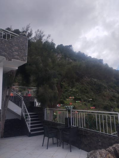 The image depicts an outdoor scene in Tétouan, Morocco, likely a terrace or patio area of a building situated on a hillside. The weather appears overcast with a cloudy sky. In the foreground, there are black chairs and a table on a tiled surface. A white railing runs along the edge of the terrace, beyond which the hillside slopes upwards. On the hillside, there are scattered groups of brightly colored chairs and tables, suggesting a recreational or dining area. Vegetation covers the hillside, including trees and shrubs. A set of stairs with white railings leads up the hill, seemingly connecting different levels of the building or outdoor space. The architecture of the building is visible on the left, featuring stone accents and white walls.