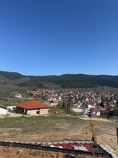 The image shows a view of Pavelsko, Bulgaria, with a clear blue sky above. A tree-covered mountain range runs parallel to the town. In the foreground is an open grassy area with a building under construction with a terracotta-colored roof. A border of dark-colored bricks and red mulch with some greenery separates the grassy area from what appears to be a lower level. A wooden structure and white rope are in the field. The town is densely populated with buildings of varying colors and heights, primarily with red-tiled roofs.