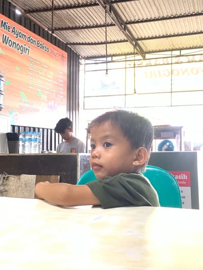 A child sits at a table in an outdoor eatery. The eatery is called "Mie Ayam dan Bakso Joyo Wonogiri", and the sign lists various "Bakso" dishes and prices in Indonesian Rupiah. The menu includes "Bakso Halus + Tetelan", "Bakso Halus Komplit + Tetelan", "Bakso Urat Jumbo + Tetelan", "Bakso Urat Jumbo Komplit + Tetelan", "Bakso Isi Daging + Tetelan", and "Bakso Campur + Tetelan." A man in a grey shirt sits at a table nearby. Bottles of drinking water are on the table. The eatery is located in Samarinda, Indonesia, as indicated by the phone number with Indonesian country code on the menu.
