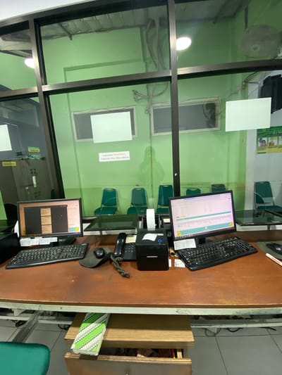 The image shows a desk with two computer monitors, two keyboards, a mouse, a telephone, and a small black printer. There are also some papers and a few books. The desk is made of wood and is located in a room with green walls. Through a glass partition, several empty green chairs can be seen, as well as a white board on the wall. There is also a large fan mounted on the ceiling.
A sign on the wall behind the chairs reads "Lakukan Identifikasi Sebelum Obat Diserahkan" which translates to "Perform Identification Before Dispensing Medication." Another sign at the bottom of the image reads "DORONG" which means "Push." The overall impression is of an office or waiting area, possibly in a healthcare facility in Margoyoso, Indonesia, given the Indonesian text. The lighting suggests it could be daytime. There are no people visible in the image, but the setup implies that work is either ongoing or has recently concluded.