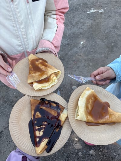 Three individuals stand on a grey, possibly asphalt or concrete, surface. Each person is holding a paper plate with a crêpe drizzled with sauce. The plate held by the person on the top right has a light brown sauce, while the plate on the bottom left has a similar light brown sauce with a smaller portion. The plate on the bottom right holds a crêpe with a dark, glossy sauce. Each person also holds a clear, plastic fork or spork. The individuals are wearing winter jackets; the person on the top right has a light blue jacket, the person on the bottom left has a pink and white jacket, and the person on the bottom right has a purple jacket. The location is unspecified, but provided information indicates the city of Nizhnie Yaki, Russia.