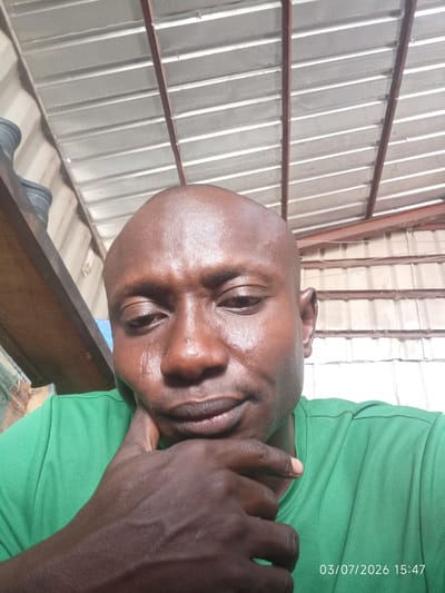A close-up, low-angle selfie shows a Black male with a shaved head, wearing a green polo shirt. He is resting his chin on his right hand, with his left hand near his face. His skin is glistening, suggesting sweat or oil. A visible tear track runs down his left cheek. The background is an industrial or workshop setting with a corrugated metal roof supported by exposed beams. To the left, a wooden shelf with what appears to be gardening pots is visible. The timestamp in the lower right corner reads "03/07/2026 15:47". The scene is likely in Bana Stream, Nigeria, given the context.