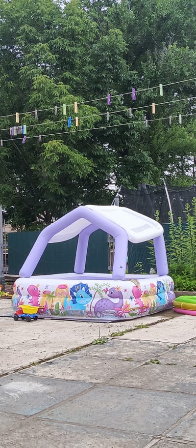 In Pogranichny, Russia, an inflatable paddling pool is set up on a paved patio area. The pool is decorated with colorful cartoon dinosaurs and features a purple inflatable canopy supported by four poles. To the left of the pool, a small yellow toy truck is visible. Behind the pool, a green wooden fence and lush green trees form the background. Clotheslines with several colorful clothespins are strung across the upper portion of the image, against the backdrop of the trees. The environment appears to be an outdoor residential area.