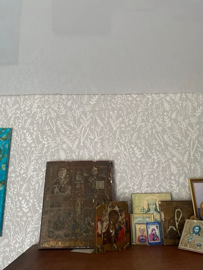 The image depicts a collection of religious icons displayed on a dark brown wooden surface, likely a shelf or dresser. The background is wallpapered with a delicate, light grey pattern of white floral and leafy motifs. To the left, a portion of a framed artwork with bright blue and green hues is visible.
The icons themselves vary in size and condition. The largest icon, positioned centrally, is dark and appears aged, with multiple figures and scenes depicted. To its right, a smaller icon shows figures of Mary and Jesus. Other icons are arranged to the right, including one with a crowned female figure and another featuring a bearded male figure. Some icons are framed.
The overall impression is that of a personal shrine or collection of religious artifacts. There are no people visible in the image, and no actions are taking place. The scene is static and orderly. The lighting appears to be natural or from an overhead source, casting soft shadows. The image quality is good, with clear details of the icons and wallpaper.
This scene is located in the city of Sakmara, Russia, within a domestic interior.