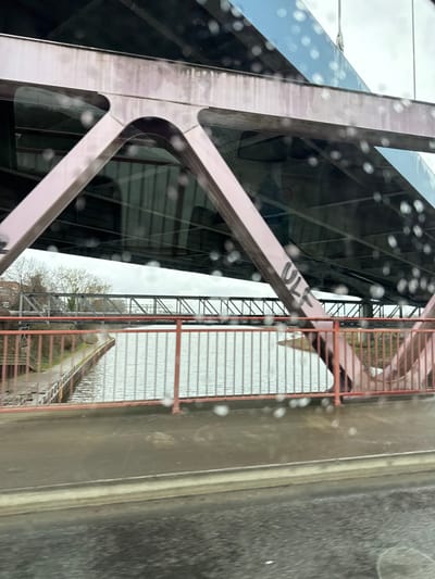 The image depicts a view from inside a vehicle looking out onto the Rhein-Herne Canal in Bottrop, Germany, specifically featuring the Rheinbabenbrücke (Rheinbaben Bridge) carrying the B224. In the immediate foreground, a blurred dark surface, likely the vehicle's dashboard or part of the road, is visible. The camera lens or window is covered in numerous raindrops, creating a speckled, out-of-focus effect across the entire image.
The prominent subject is the large, weathered reddish-brown metal truss structure of the Rheinbabenbrücke, which arches over the canal. Its robust beams and supports are visible from below the bridge deck. Black graffiti, spelling "ULF", is visible on one of the vertical beams on the right side of the main bridge structure. Below the main bridge deck, a red-orange painted metal railing with vertical bars runs along the edge of the bridge, overlooking the canal.
Below the Rheinbabenbrücke, the grey, rippled surface of the Rhein-Herne Canal flows. The left bank of the canal features a paved path running alongside a concrete or stone retaining wall, partially obscured by more red-orange vertical railings. On the far bank, sparse, leafless trees are visible against an overcast, light grey sky. In the mid-ground, spanning the canal parallel to the Rheinbabenbrücke, is another, darker-colored metal truss bridge, likely a railway or industrial bridge, with a more intricate lattice work design. No people or active interactions are visible in the scene. The overall environment suggests an overcast, damp, possibly rainy day.